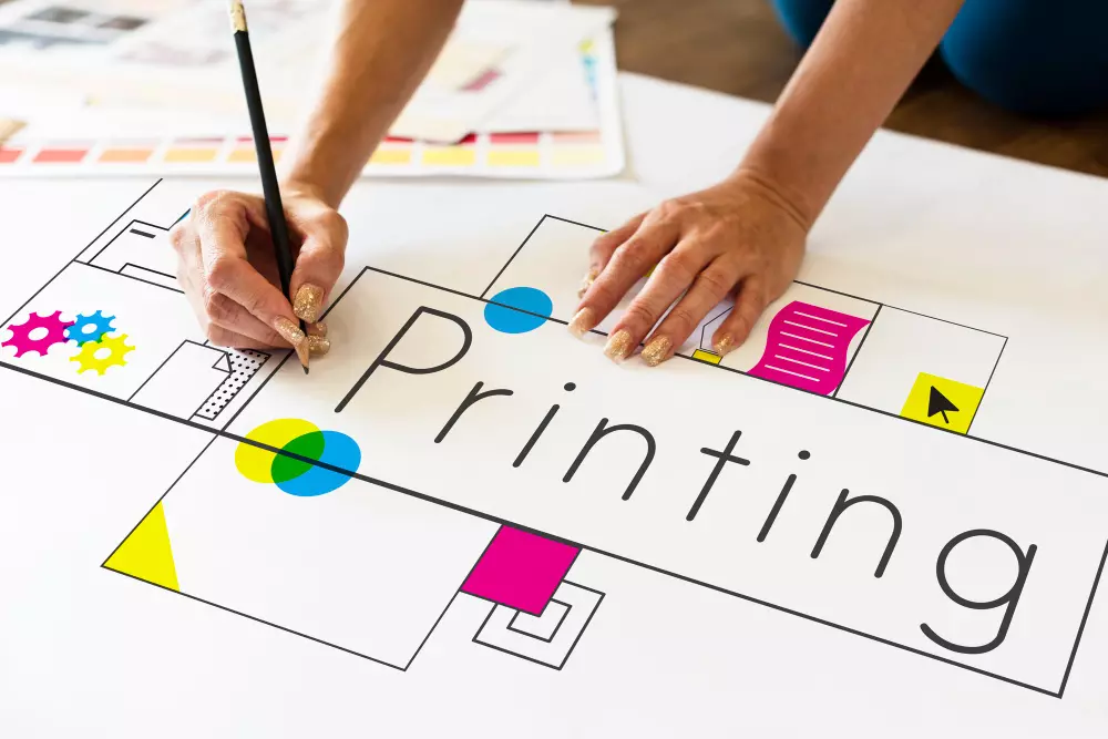 La imagen muestra las manos de una persona trabajando en un boceto para un diseño de impresión. El papel sobre la mesa tiene la palabra "Printing" en el centro con iconos gráficos y paletas de colores a su alrededor, sugiriendo un diseño en proceso para servicios de impresión. La persona está usando un lápiz negro y tiene las uñas pintadas con brillo, indicando una fase de planificación y creatividad en el desarrollo de un producto de impresión.