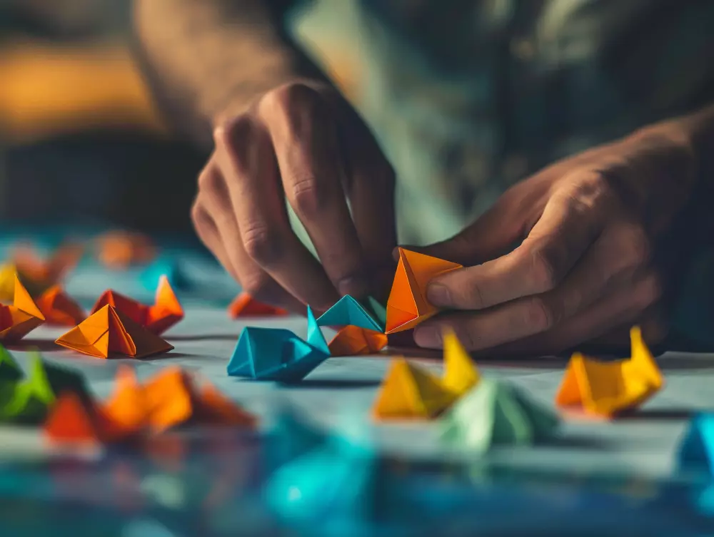 La imagen muestra las manos de una persona manipulando coloridos barcos de papel origami sobre una mesa. Los barquitos, en tonos vibrantes de naranja, azul, verde y amarillo, están esparcidos por la superficie, creando una escena artística y meticulosa. Este detalle evoca un sentido de creatividad y precisión, destacando el enfoque y la delicadeza involucrados en el arte del origami.