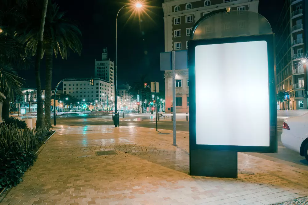 La imagen muestra una luminosa pantalla publicitaria en una calle urbana durante la noche. El anuncio está vacío, listo para ser utilizado, y se encuentra en una acera bien iluminada, con edificios y árboles de fondo. Las luces de la calle y los automóviles crean un ambiente vibrante y dinámico, destacando el potencial de la publicidad exterior para captar la atención de los transeúntes en un entorno nocturno.