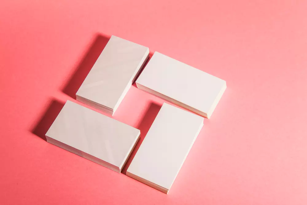 La imagen muestra cuatro pilas de tarjetas de visita en blanco, dispuestas de manera ordenada sobre un fondo rosa. Las tarjetas están alineadas en una disposición geométrica, destacando su tamaño y forma estándar. Este diseño minimalista resalta la versatilidad y el potencial creativo que ofrecen las tarjetas de visita para reflejar la identidad corporativa y el branding personal.