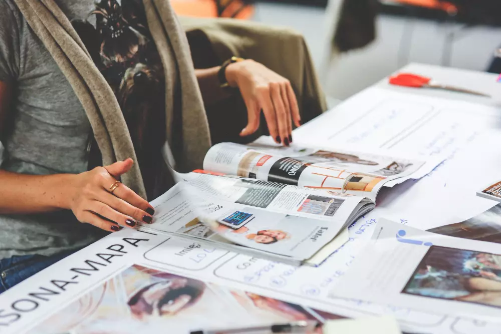 Persona revisando un catálogo impreso en una mesa de trabajo, rodeada de documentos y materiales de diseño, enfocada en la planificación y creación de contenidos visuales.