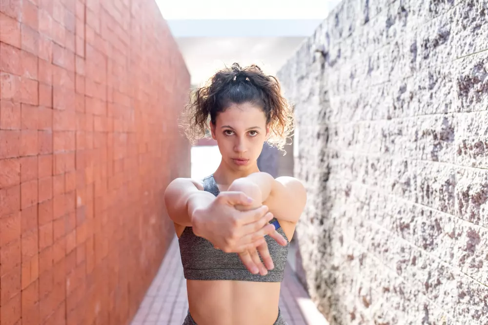 Una mujer joven en ropa deportiva estirando los brazos frente a ella, en un pasillo al aire libre con paredes de ladrillo rojo y gris a los lados. La expresión determinada en su rostro refleja concentración y enfoque en su rutina de ejercicio.