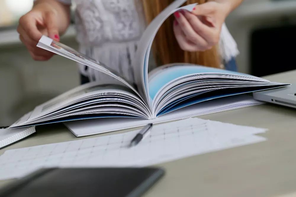 Persona hojeando una revista en una mesa de trabajo, rodeada de documentos y una computadora portátil, enfocada en la revisión o planificación de contenido editorial.