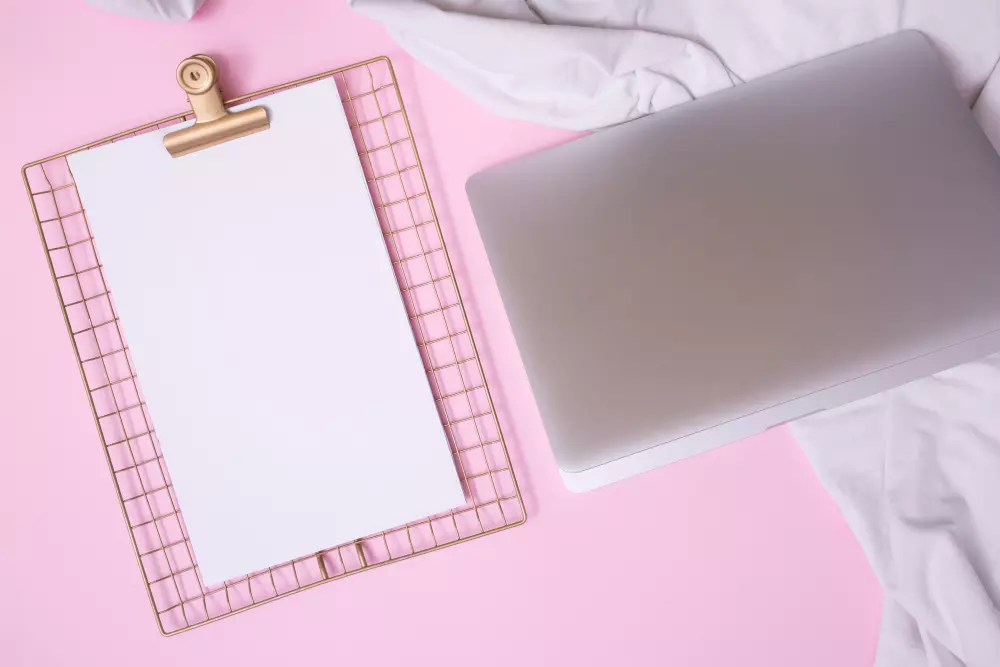 Escritorio con una computadora portátil plateada y una hoja de papel en una rejilla dorada sobre un fondo rosa, representando la preparación de documentos de diseño gráfico.