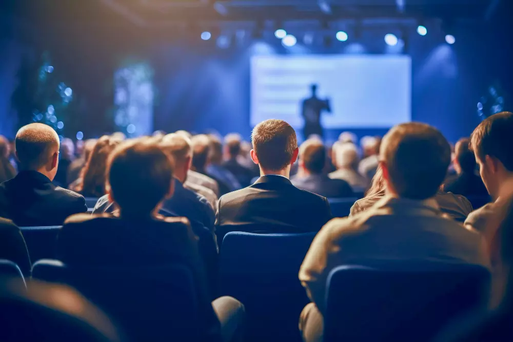 Una audiencia de personas sentadas en una sala de conferencias, enfocadas en un presentador que habla desde el escenario iluminado. La atmósfera es profesional y está llena de energía, con luces azules que añaden un toque dinámico al evento.