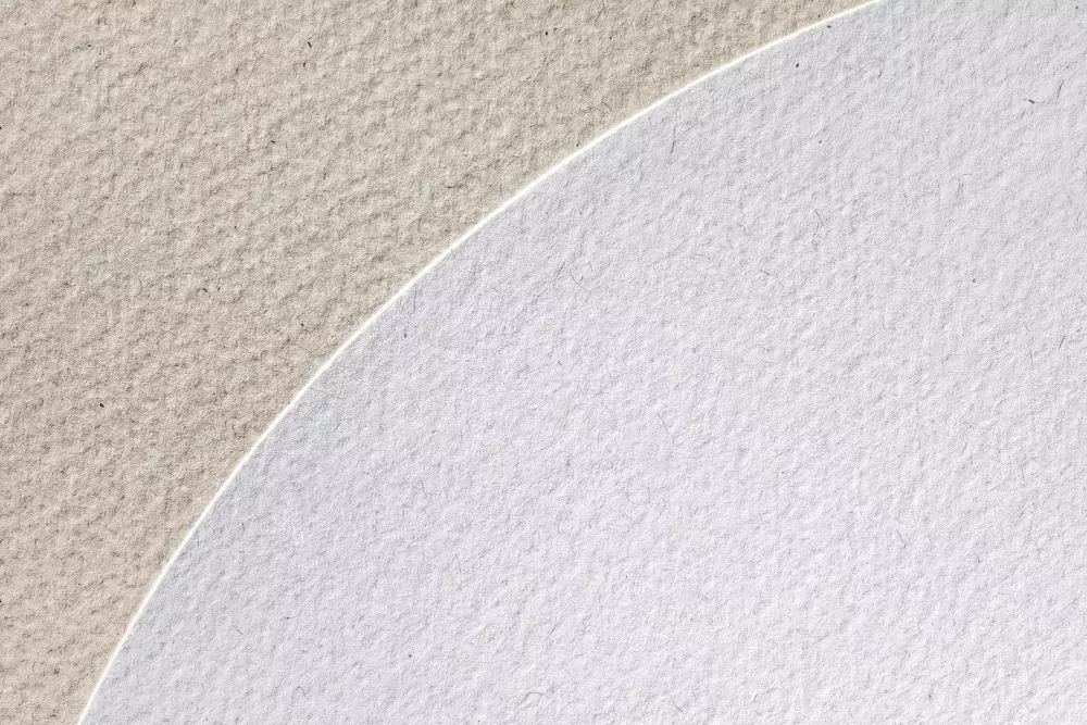 Primer plano de dos tipos de papel con texturas diferentes, una superficie ligeramente rugosa en tonos beige y otra en blanco, representando la variedad de acabados disponibles para materiales impresos y cómo estos pueden influir en la estética y la sensación táctil del producto final.