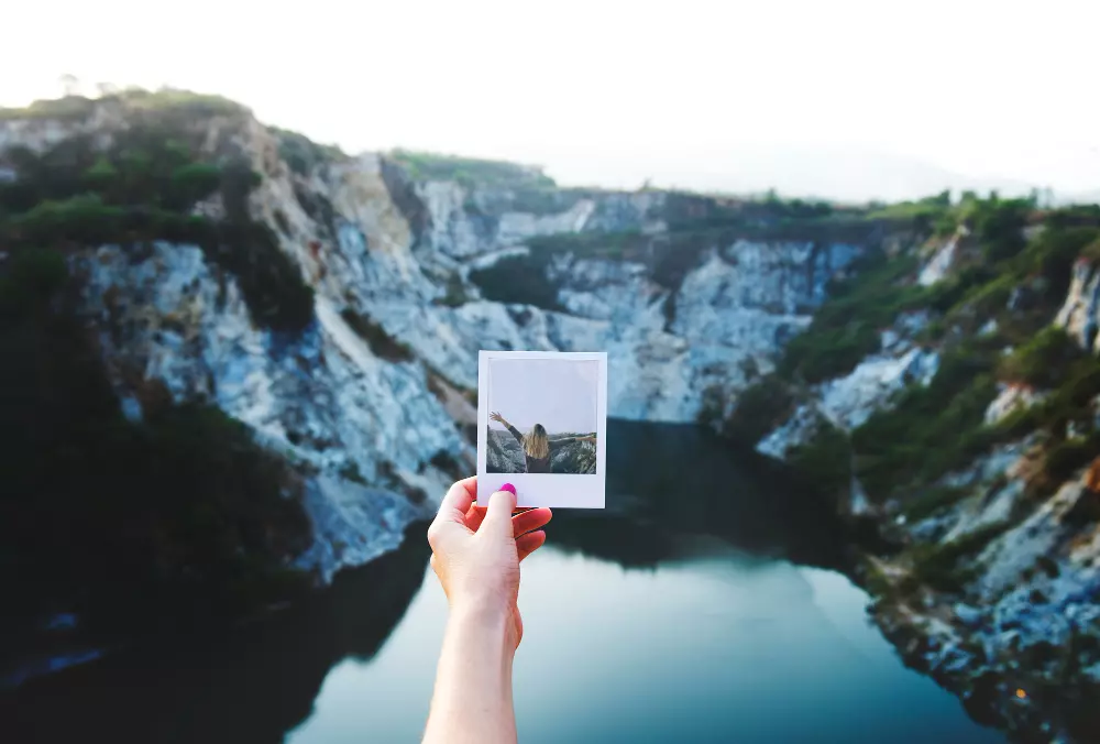 Mano sosteniendo una fotografía instantánea frente a un paisaje montañoso con un lago, destacando la conexión entre la captura de imágenes y su impresión en formato físico.