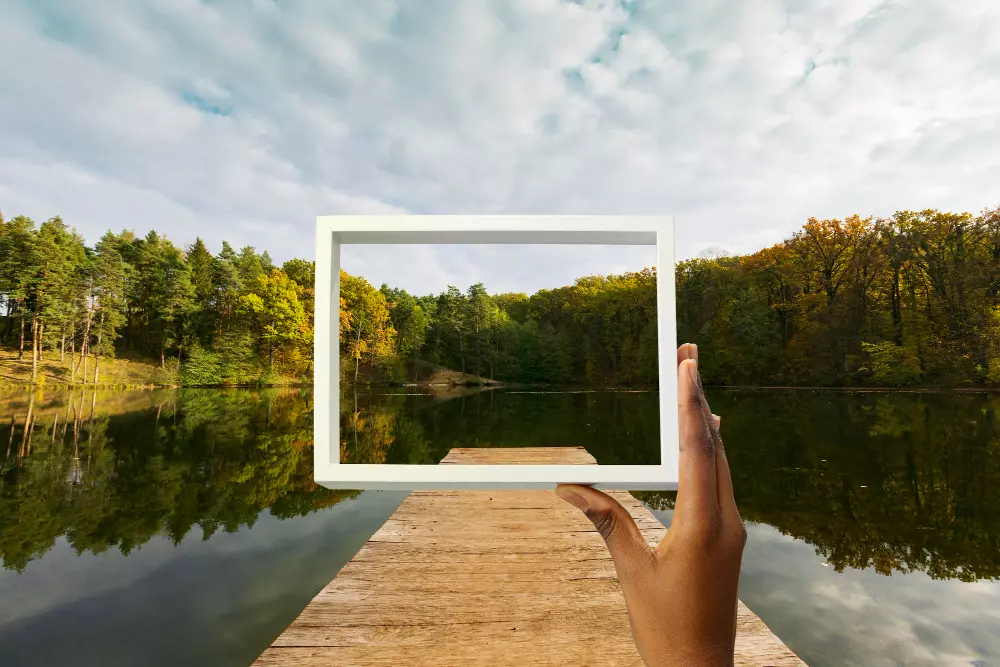 Mano sosteniendo un marco vacío con un paisaje de lago y bosque en el fondo, simbolizando la importancia de ajustar la resolución de las imágenes para obtener una impresión nítida y clara, destacando cómo diferentes resoluciones afectan la calidad de la impresión en distintos entornos y medios.