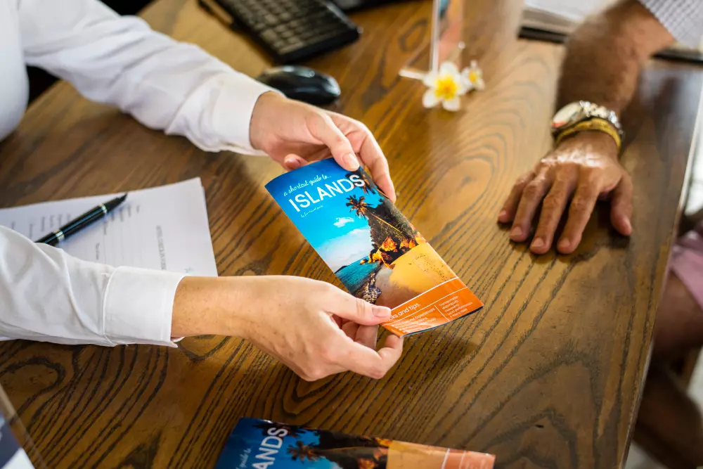 Una persona entrega un folleto turístico, un ejemplo de crear flyers personalizados, con un diseño colorido sobre islas tropicales, en una mesa de oficina donde hay documentos y un bolígrafo.